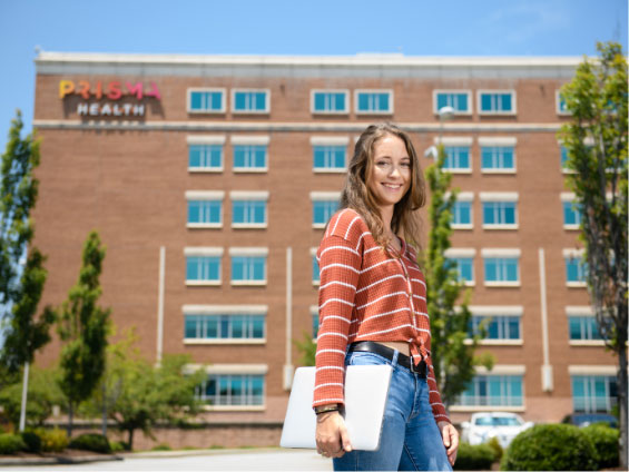 Emily stands outside of a brick hospital, smiling and holding her laptop, wearing jeans and an orange striped shirt.