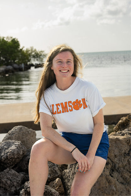 Student Erin Griffin wears a white, short-sleeve Clemson shirt and is smiling and sitting on rocks with a pier and water in the background. 