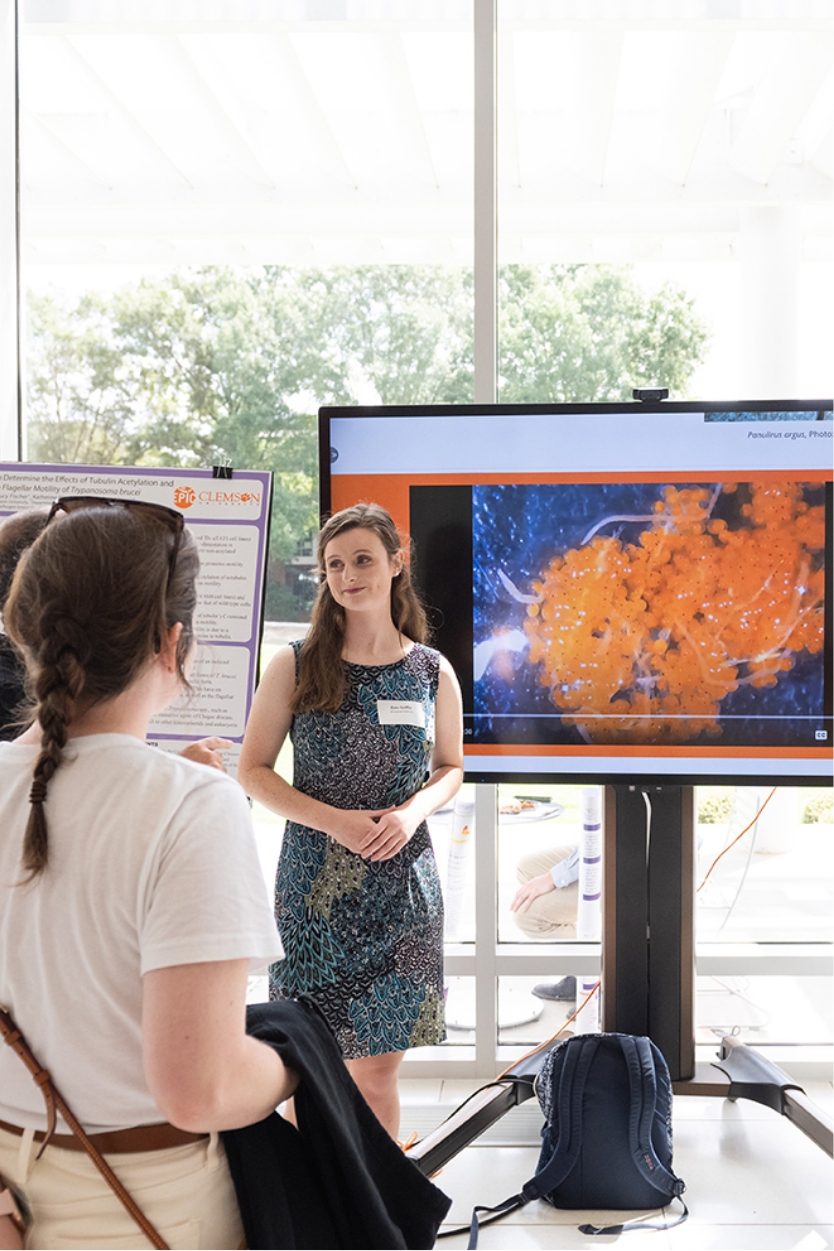 Student Erin Griffin wears a dark, flowered dress as she presents marine research on a large screen to a group of onlookers, whose backs are to the camera. They are inside, but a large window behind Erin has a canopy of trees visible in the background. 