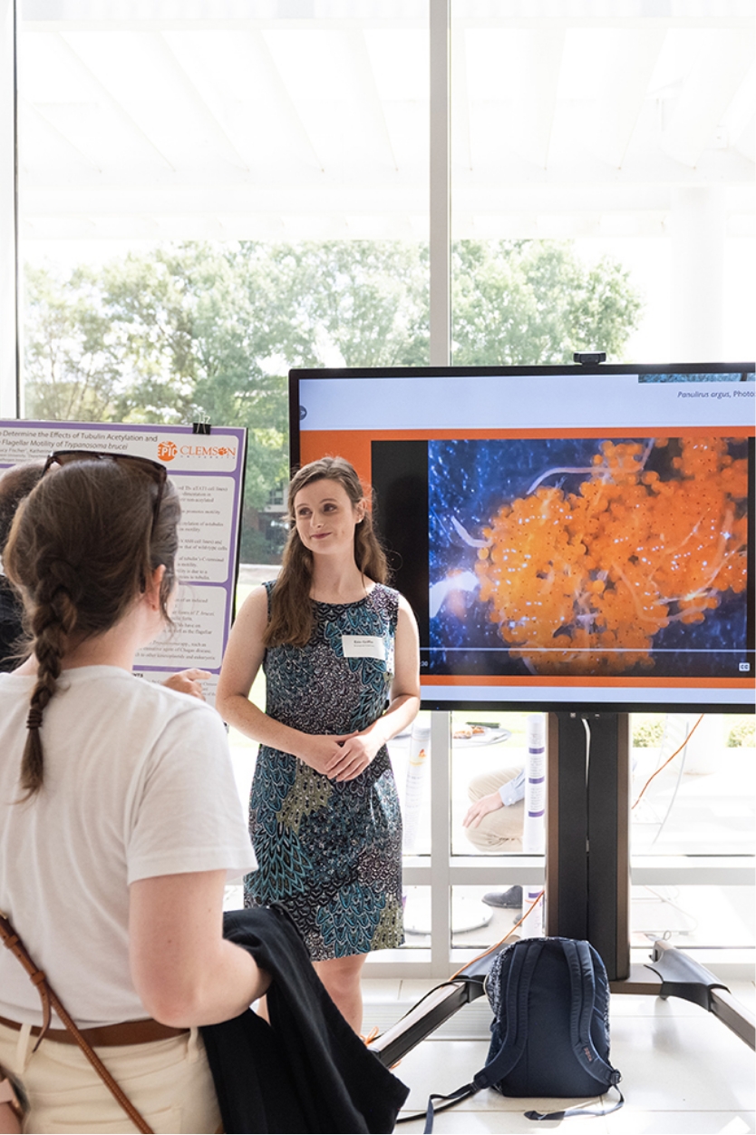 Student Erin Griffin wears a dark, flowered dress as she presents marine research on a large screen to a group of onlookers, whose backs are to the camera. They are inside, but a large window behind Erin has a canopy of trees visible in the background. 