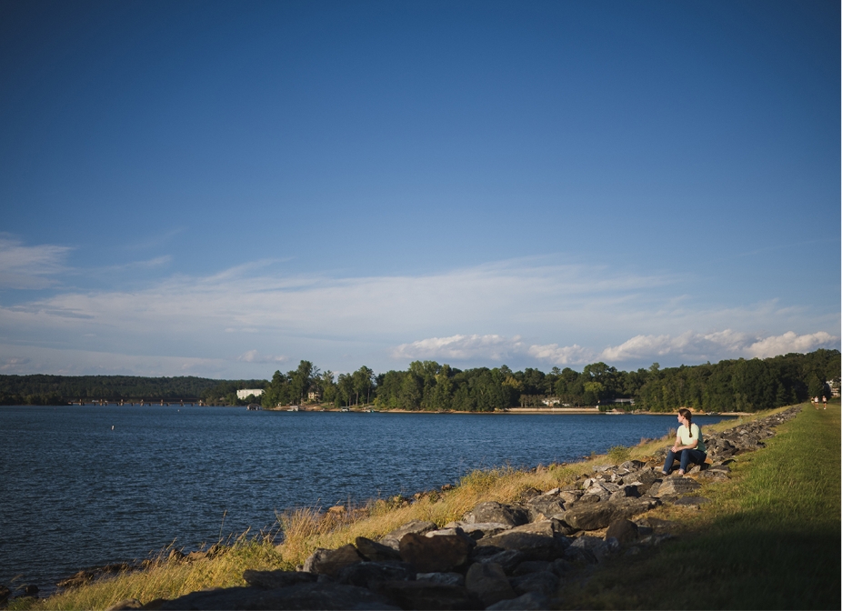 A wide landscape of Lake Hartwell shows the water, rocky shoreline and trees.  Student Erin Griffin can be seen distantly, seated on the rocks overlooking the lake. 