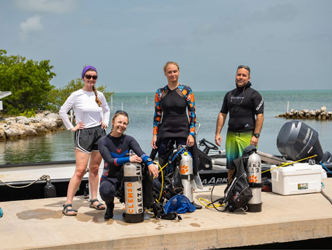 Professor Antonio Baeza and his team of student researchers are standing on a pier looking at the camera, holding air tanks and diving gear, with a boat and water in the background.