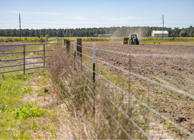 A fence seperates two fields. On the right field a tractor is driving and there is a building in the background.