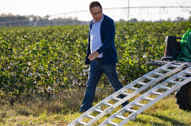 Joe is walking in front of a field of crops preparing to unload equipment off of a four wheeler.