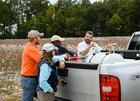 Kendall Kirk and three students are in a field of crops standing next to a pickup truck. The three students are leaning against the bed of the truck as Kendall Kirk explains something to them.