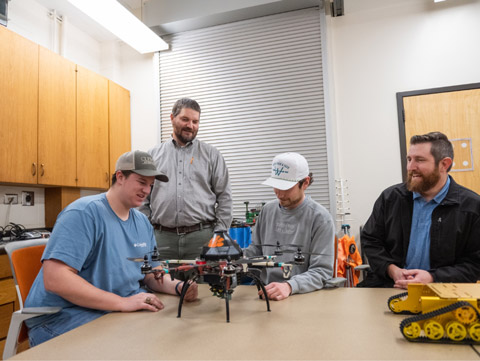 Three students sit at a table with Kendall Kirk. They are all inspecting a drone inside of a office.