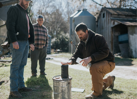 At a farm Kendall Kirk is kneeling down on the ground and showing two farmers how a piece of technology works.