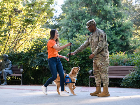 Marena hands off a service dog to a man in military uniform in front of a pair of park benches and some bushes.