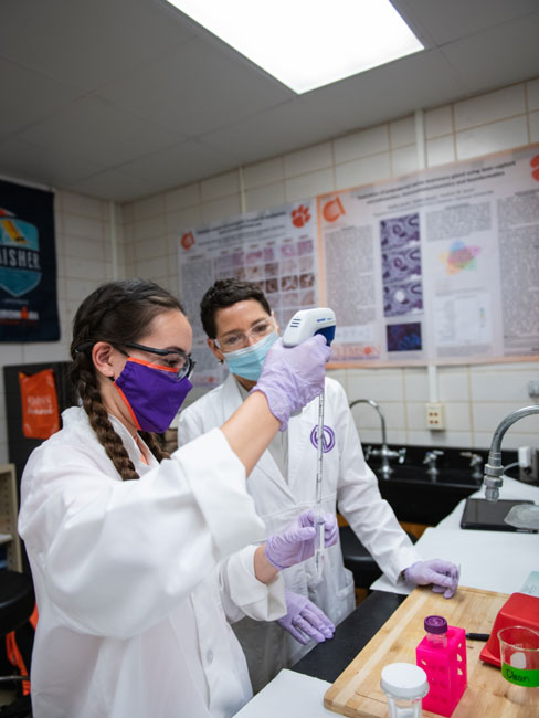 Marena works with a colleague in a lab to observe and measure a substance in a test tube.