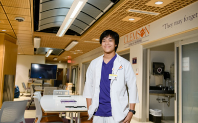 Nam stands, smiling with hands in his pockets in a brightly lit nursing simulation lab wearing white and purple Clemson scrubs.