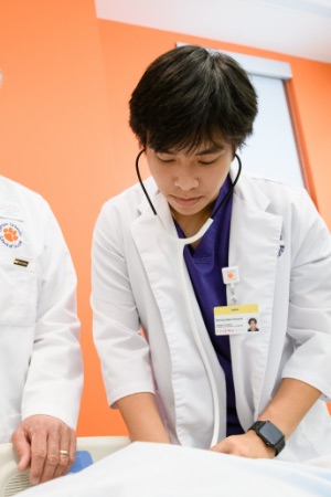 Nam takes the pulse of a human simulator in the lab by his professor, using a stethoscope, wearing purple and white scrubs.