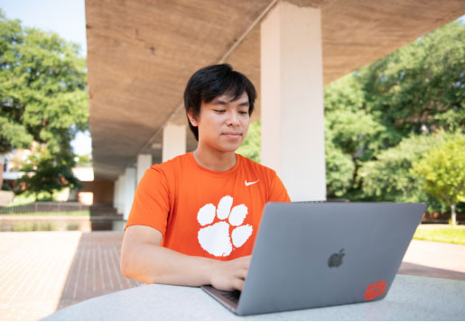 Nam, in an orange shirt with a Tiger Paw, works on his laptop at a table in the shade under a bridge with trees in the background.