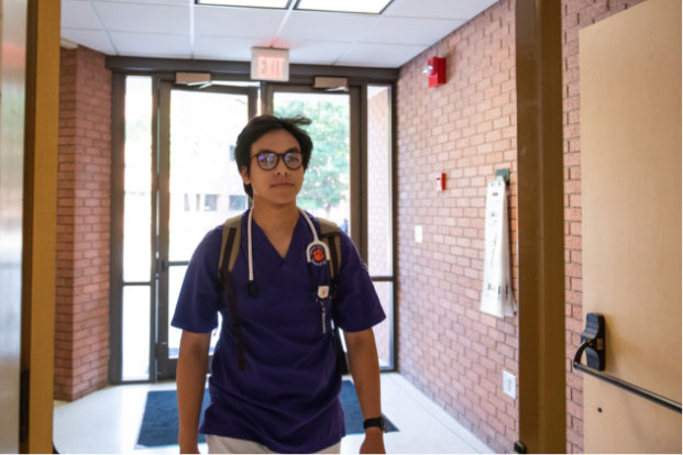 Nam walks into Edwards Hall, wearing purple and white scrubs, a stethoscope, a backpack and glasses.