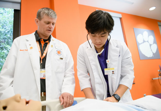 Nam uses a stethoscope to gauge the pulse of a human simulator in the nursing lab while his professor stands beside him.