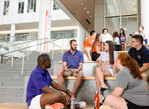Hunter sits on steps with a group of Clemson students and has a conversation with them. Another group of students are sitting behind him having their own conversation.