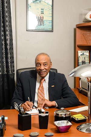 Roy Jones looks at the camera while writing a letter at his desk, wearing a black suit and orange tie.