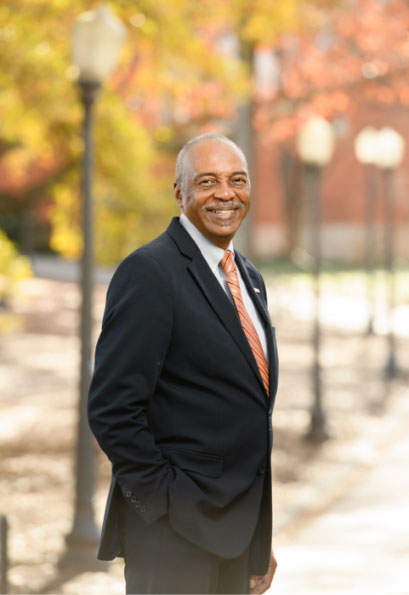 Roy stands smiling on a sidewalk lined by bright colored trees in fall, wearing a black suit and orange tie.