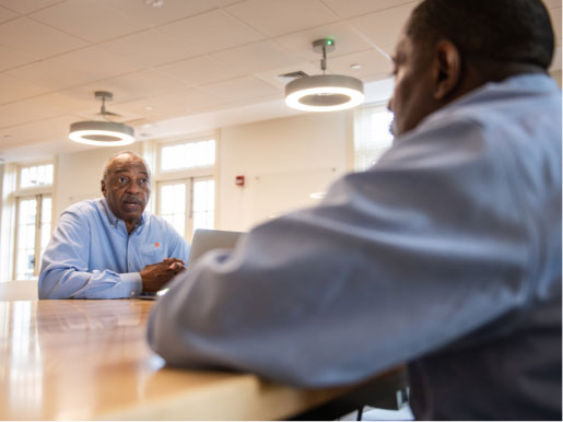 Roy sits at a wood table and talks with his colleague Mark. Both men wear light blue Call Me MISTER® button downs.