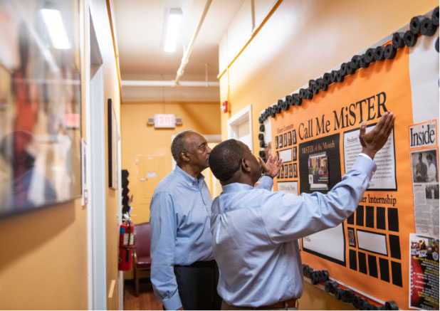 Roy listens as his colleague Mark discusses the Call Me MISTER results posted to a decorative board in their office space.