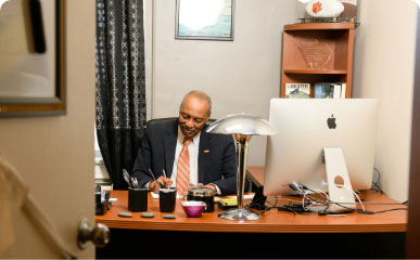 Roy Jones sits and writes a letter at a wood desk in his office wearing a black suit and orange tie.