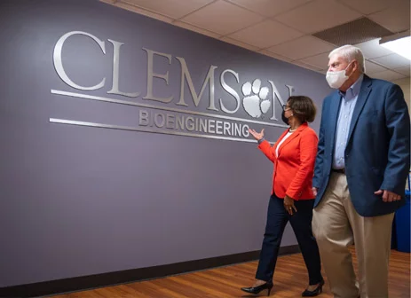 Vanessa gestures at a Clemson Bioengineering sign while walking with a companion in a hallway