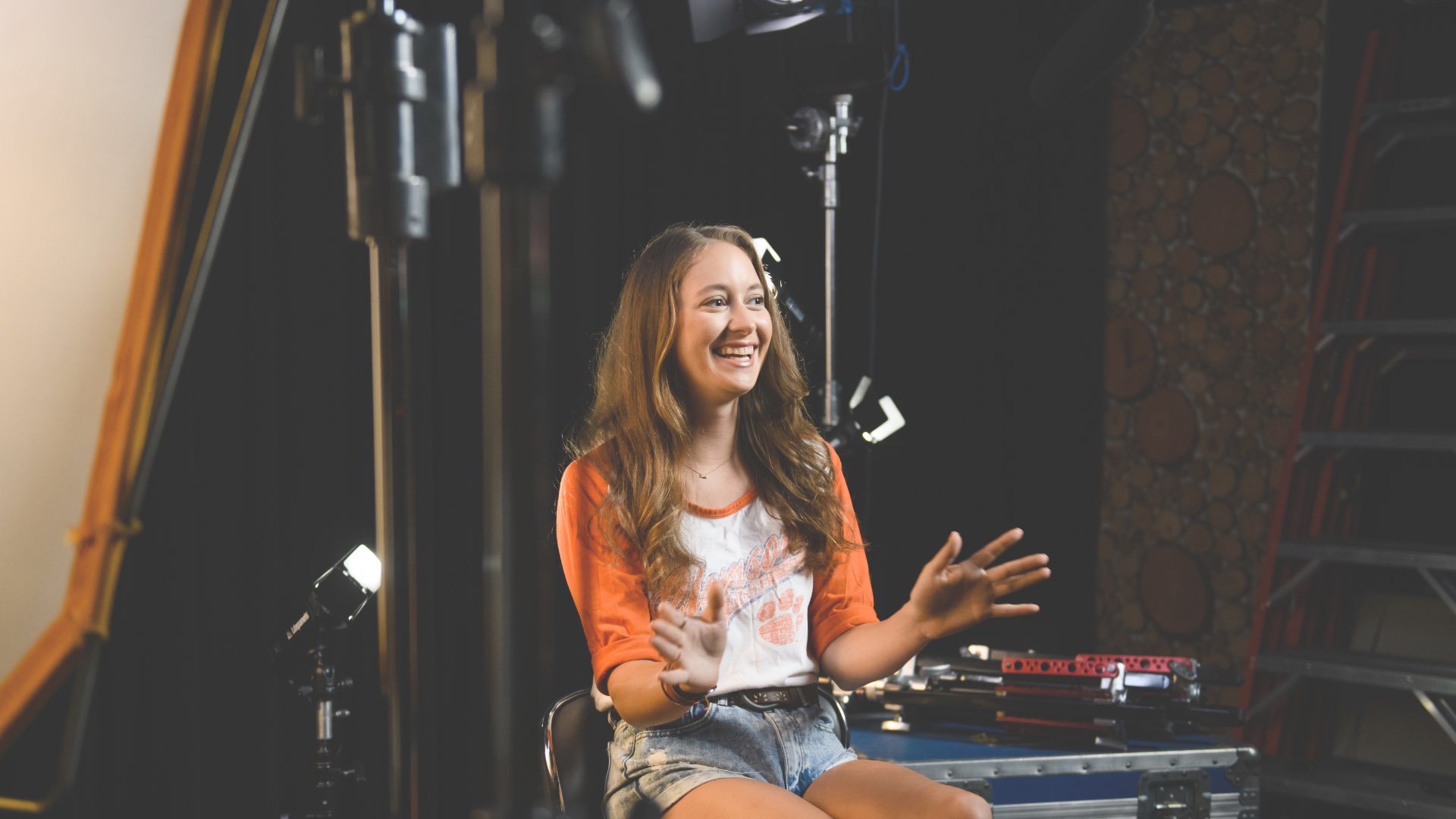 Emily sits in a backstage area, smiling and gesturing with her hands.'Watch Emily's Story' and the play symbol are beside her.