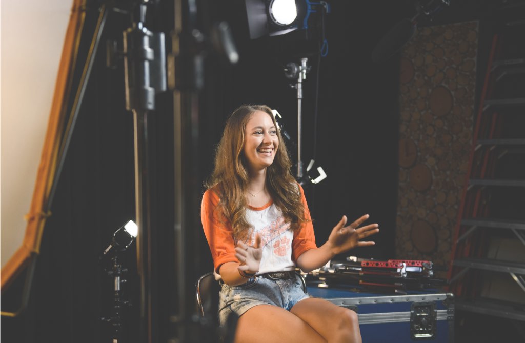 Emily sits in a backstage area, smiling and gesturing with her hands.'Watch Emily's Story' and the play symbol are beside her.