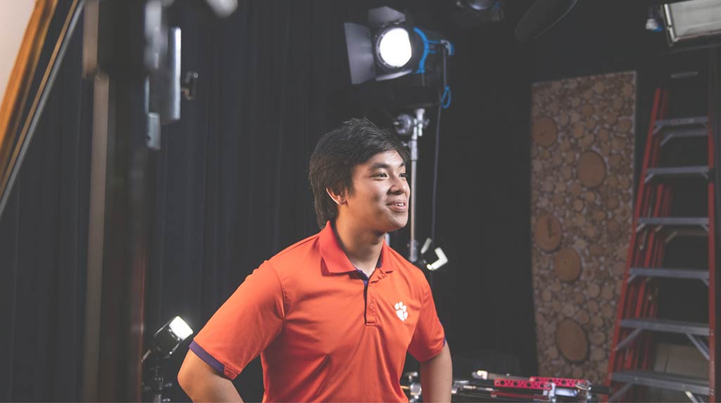 Nam sits in a backstage video area wearing an orange Clemson polo and talking. 'Watch Nam's Story' and the play symbol hover beside him.