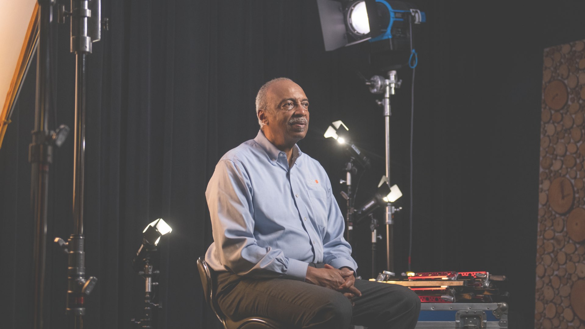 Roy sits in a backstage video area, looking thoughtfully ahead of him. 'Watch Roy's Story' and the play symbol hover beside him.