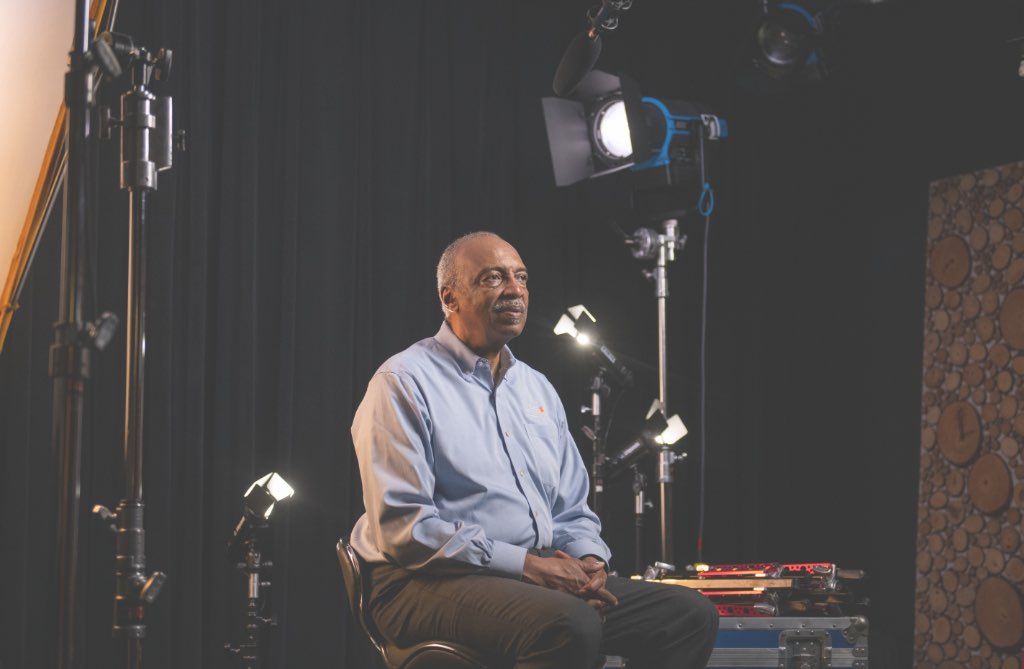 Roy sits in a backstage video area, looking thoughtfully ahead of him. 'Watch Roy's Story' and the play symbol hover beside him.