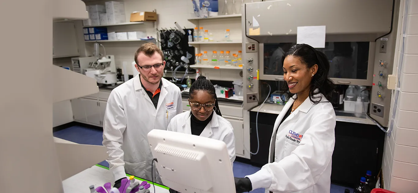 Two female researchers and one male researcher in white lab coats examine a monitor with lab equipment in the background