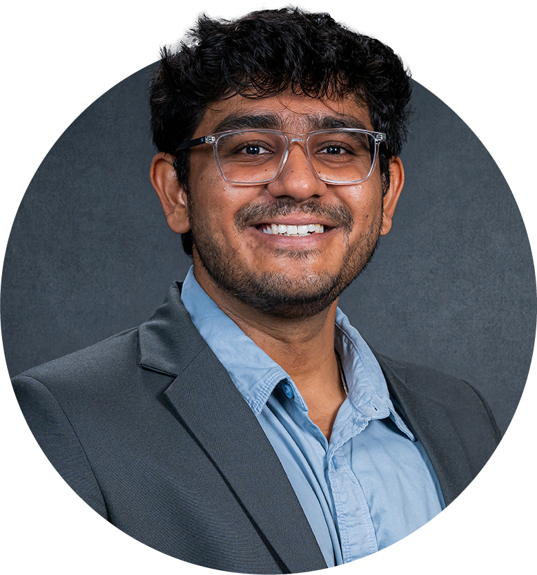 A male graduate student wearing a gray blazer, light blue button-down shirt and glasses poses for a headshot.