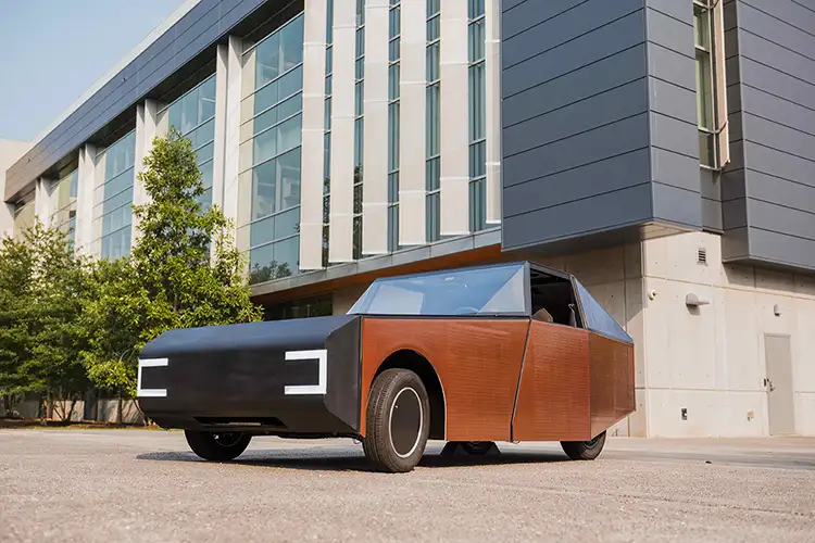 The Deep Orange 17 prototype vehicle, featuring a futuristic, angular design with a black front, wood-toned sides and blue-tinted windows, is parked outside a modern glass-and-concrete building on a sunny day.