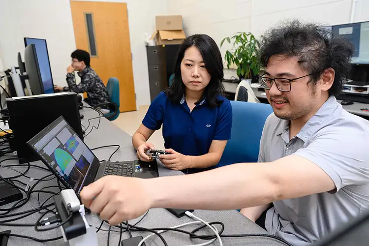 A female professor and a male student sit together in an office in front of a computer workstation, examining data on a laptop while handling wired electronic equipment.