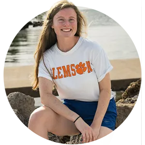 A female student wearing blue running shorts and a white T-shirt that says “Clemson” in orange poses on a rocky ledge in front of a body of water.