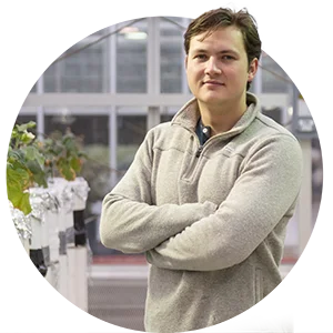 A male student wearing a light gray fleece pullover with his arms folded stands in an on-campus greenhouse research facility.