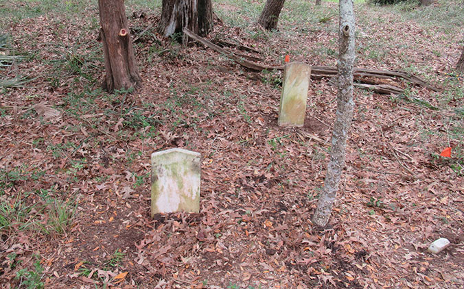 Two basic headstones with spots of moss standing among a few trees and bright orange stake flags nearby.