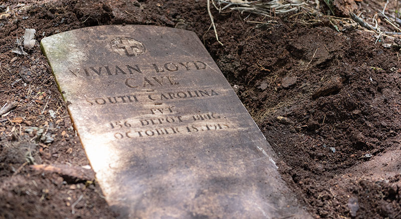 Headstone laying down surrounded by freshly dug mud. The headstone displays an etched cross and lettering that appear to say 'Vivian Loyd Cave South Carolina 156 Depot October 18, 1918.' Some text is not legible.