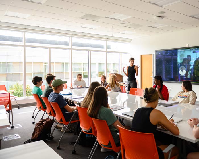 Students sit around a large table while a female instructor lectures at a whiteboard