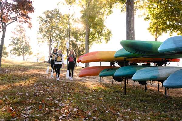 three students in lifejackets walk alongside canoes and kayaks
