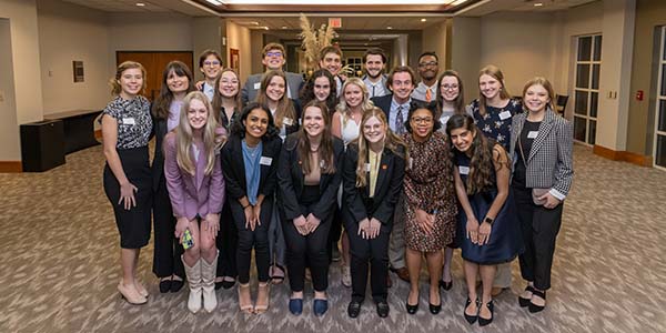 A group of students standing closely together and smiling for the camera in the lobby of the Madren Center on campus. 