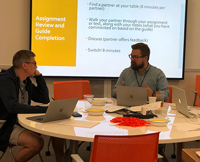 Two men having a discussion with a projector displaying Assignment Review in the background