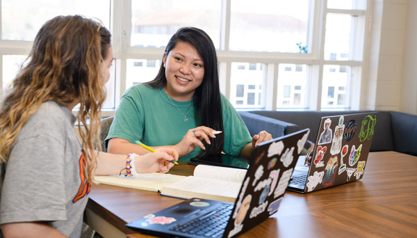 Two female students discuss at a table over their computers and notebooks