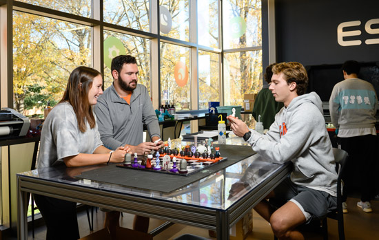 two students sit with a faculty member around a table with a 3d printed chess board sitting on top inside a classroom