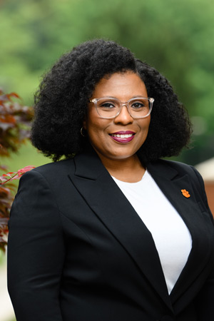 A dark haired woman in a blazer and a tiger paw pin smiles with greenery in the background.