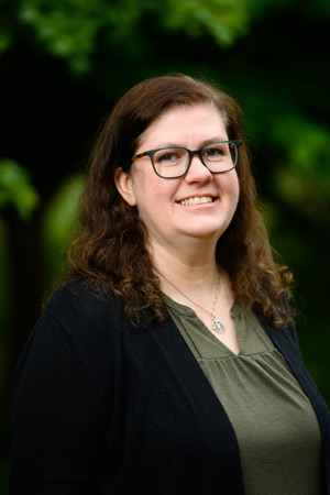 A woman with brown hair smiles with greenery in the background.