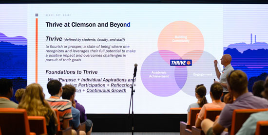 A male staff member gives a presentation to a large group of freshman on a wall sized digital screen. 