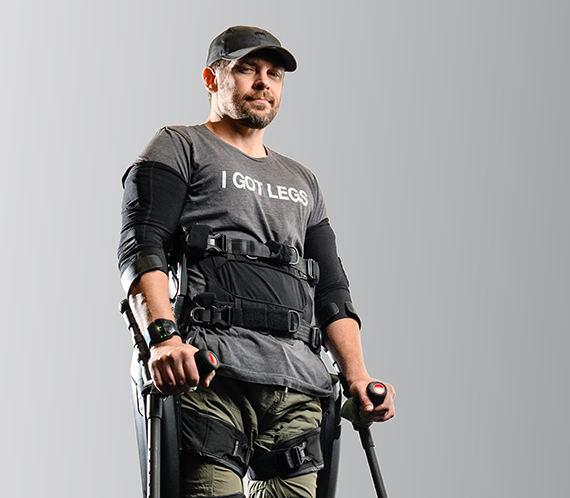 Adam Gorlitsky wearing a shirt which says I got legs, stands assisted by leg, torso and arm braces.