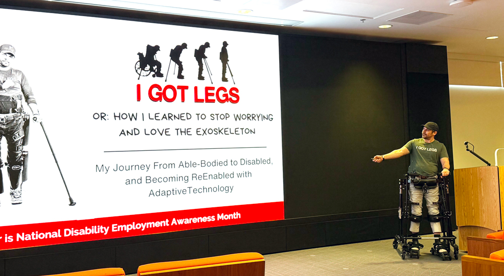 Adam Gorlitsky stands with assistance from an exoskeleton and points to the title slide for his presentation: I got legs, how I learned to stop worrying and love the exoskeleton.