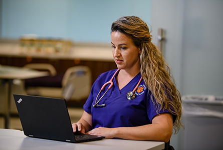 Lael Day wearing a stethescope and a purple nursing uniform with orange tiger paw working on a laptop.